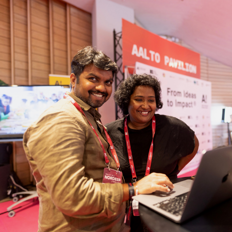 Two people smiling, one of them looking at the camera, the other one looking at the laptop in front of them
