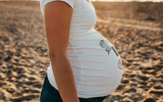 A close-up of a pregnant person wearing a white T-shirt, standing on a sandy beach at sunset.