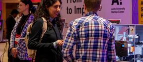 People talk at a tech fair booth, one with a backpack, another in blue checked shirt, machines beside them