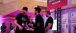 People talk at an Aalto University pavilion stand indoors with pink lighting and banners.