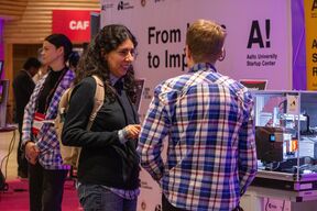 People talk at a tech fair booth, one with a backpack, another in blue checked shirt, machines beside them