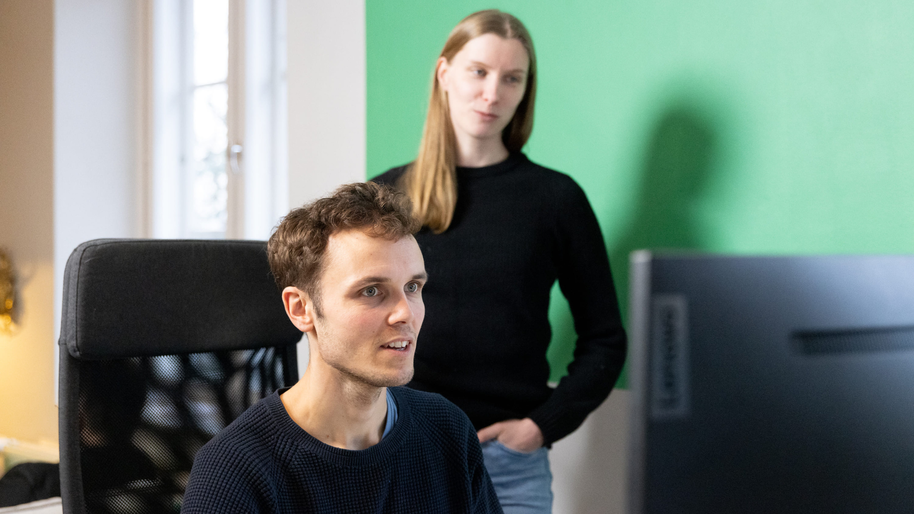 Two people from the KOKO Forest team looking at a laptop screen. A male presenting person sitting down, and a female presented one behind him standing up