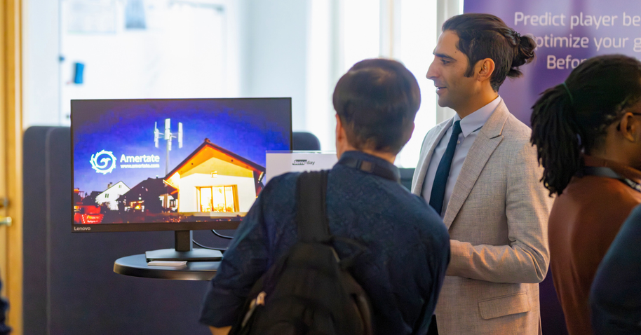 A startup founder explaining their project to an interested Demo Day attendee. In the back, a screen with promotional images for the startup.